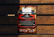 a red and black typewriter sitting on top of a wooden bench
