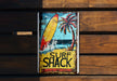 a surf shack sign hanging on a wooden wall