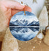 a hand holding a round ornament with a mountain scene on it