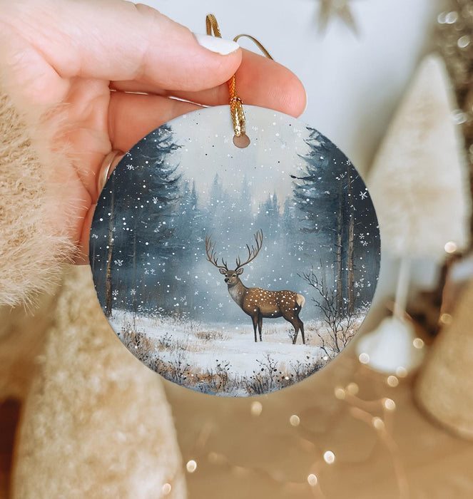 a hand holding a christmas ornament with a deer in a snowy forest