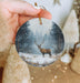 a hand holding a christmas ornament with a deer in a snowy forest