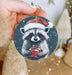 a hand holding a christmas ornament with a raccoon wearing a santa