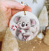 a hand holding a christmas ornament with a koala holding a candy cane