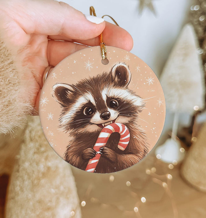 a hand holding a christmas ornament with a raccoon holding a candy
