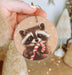 a hand holding a christmas ornament with a raccoon holding a candy