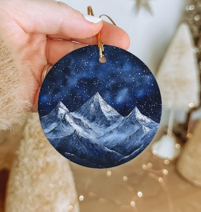 a hand holding a round ornament with a mountain scene painted on it