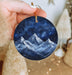 a hand holding a round ornament with a mountain scene painted on it