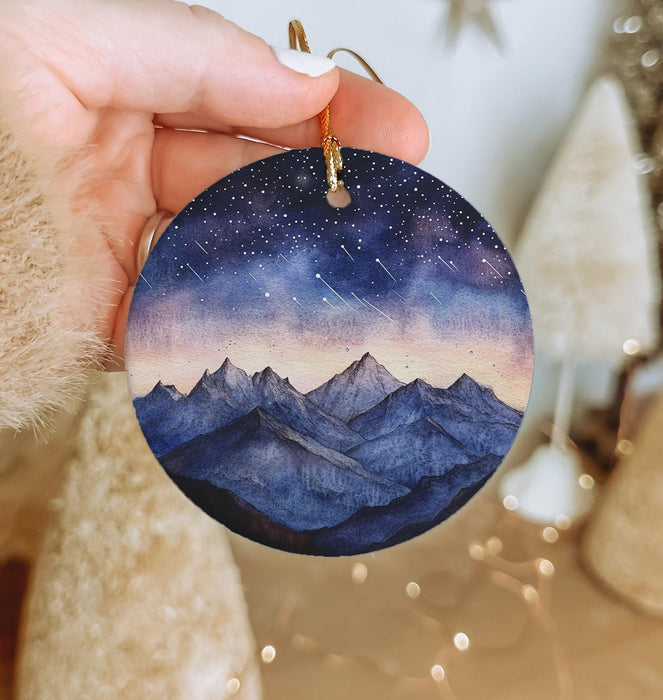 a hand holding a round ornament with a mountain scene painted on it