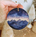 a hand holding a round ornament with a mountain scene painted on it