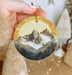 a hand holding a round ornament with a mountain scene on it