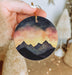 a hand holding a round ornament with a painting of mountains