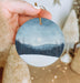 a hand holding a round ornament with a landscape painted on it