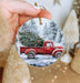 a hand holding a christmas ornament with a red truck carrying a christmas tree