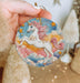 a hand holding a round ornament with a unicorn on it