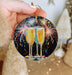a hand holding a christmas ornament with two glasses of champagne