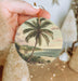 a person holding a christmas ornament with a painting of a palm tree
