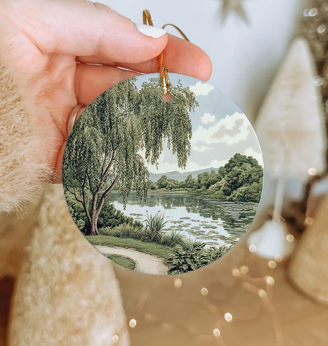 a hand holding a christmas ornament with a painting of a lake