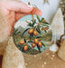 a hand holding a round ornament with a painting of oranges on it
