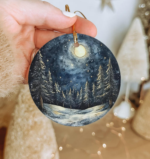 a hand holding a christmas ornament with a snowy night scene on it
