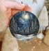 a hand holding a christmas ornament with a snowy night scene on it