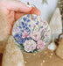 a hand holding a floral ornament in front of a christmas tree