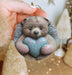 a hand holding a ceramic ornament with a teddy bear holding a heart