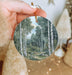 a hand holding a round ornament with a painting of a forest