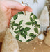 a hand holding a round ornament with green leaves on it