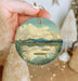 a hand holding a round ornament with a lake scene on it