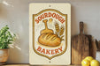 a bakery sign with a bread loaf and wheat stalks, placed on a wooden shelf next to kitchen utensils and a potted plant.