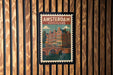 A vintage-style postage stamp depicting a cityscape of Amsterdam, Netherlands, with a bridge and canal in the background.