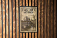 A vintage-style poster of Cardiff, Wales, featuring a castle and a bridge, is displayed on a wooden wall.