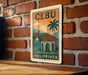 a vintage-style sign with the text "Cebu Philippines" and an illustration of a tropical beach scene with palm trees, a bridge, and a church.