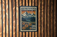 A colorful postage stamp from Colorado, featuring a mountain landscape with flowers, is displayed on a wooden wall.