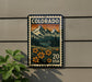 A colorful postage stamp depicting a mountain landscape with flowers, mounted on a wall next to a plant.