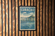 A vintage postage stamp featuring a cityscape with a river and buildings, hanging on a wooden wall.