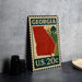 A metal sign with a red state of Georgia on it, featuring a green border and the text "GEORGIA U.S. 20c".