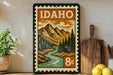 A framed vintage postage stamp depicting a mountain landscape with a river, surrounded by pine trees, is displayed on a shelf next to a wooden cutting board and some lemons.