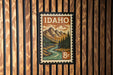 A framed postage stamp depicting a scenic mountain landscape with a river, hanging on a wooden wall.