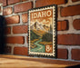 A vintage-style postage stamp from Idaho, featuring a mountain landscape with a river, is displayed on a brick wall.