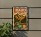A vintage Idaho postage stamp is displayed on a wall, featuring a mountain landscape with a river and pine trees.