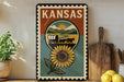 A vintage-style metal sign with the word "KANSAS" prominently displayed, featuring a sunflower and a wagon in the center. The sign is placed on a wooden shelf, with a potted plant and a wooden cutting board nearby.