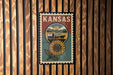 A vintage-style poster of Kansas, featuring a sunflower and a wagon, is displayed on a wooden wall.