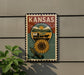 A decorative metal sign with the word "KANSAS" on it, featuring a sunflower and a wagon on a dirt road.