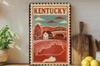 A vintage-style metal sign with a red border and the word "Kentucky" prominently displayed, depicting a rural landscape with a house and trees. The sign is placed on a wooden shelf, with a potted plant to the left and a wooden cutting board to the right.