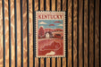 A vintage-style metal sign with the word "Kentucky" prominently displayed, featuring a rural landscape with a house and trees.