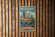 A vintage-style postage stamp from Melbourne, Australia, featuring a cityscape and a bridge, is displayed on a wooden wall.