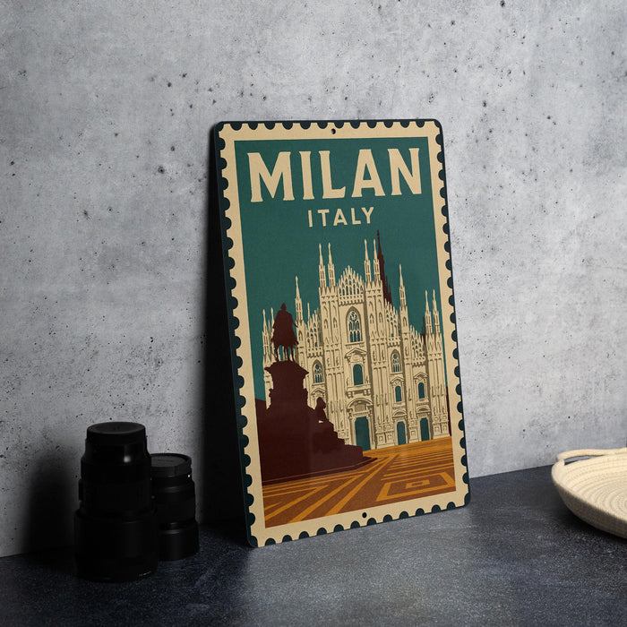 A vintage-style metal sign with the text "MILAN ITALY" and an illustration of the Milan Cathedral, placed on a gray surface next to some black camera lenses.
