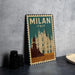 A vintage-style metal sign with the text "MILAN ITALY" and an illustration of the Milan Cathedral, placed on a gray surface next to some black camera lenses.