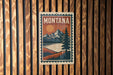 A vintage-style metal sign with the word "MONTANA" prominently displayed, featuring a mountain landscape with a pine tree and a lake.